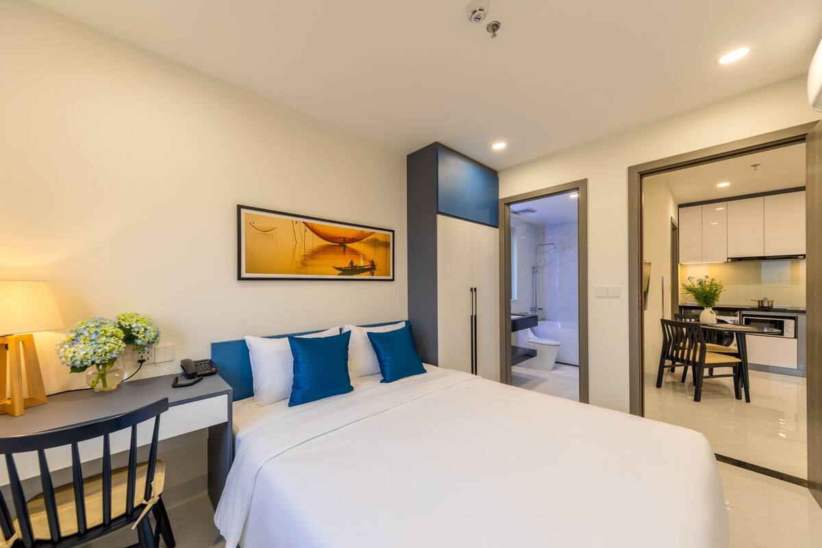 A modern hotel room with a bed, desk, and kitchenette.