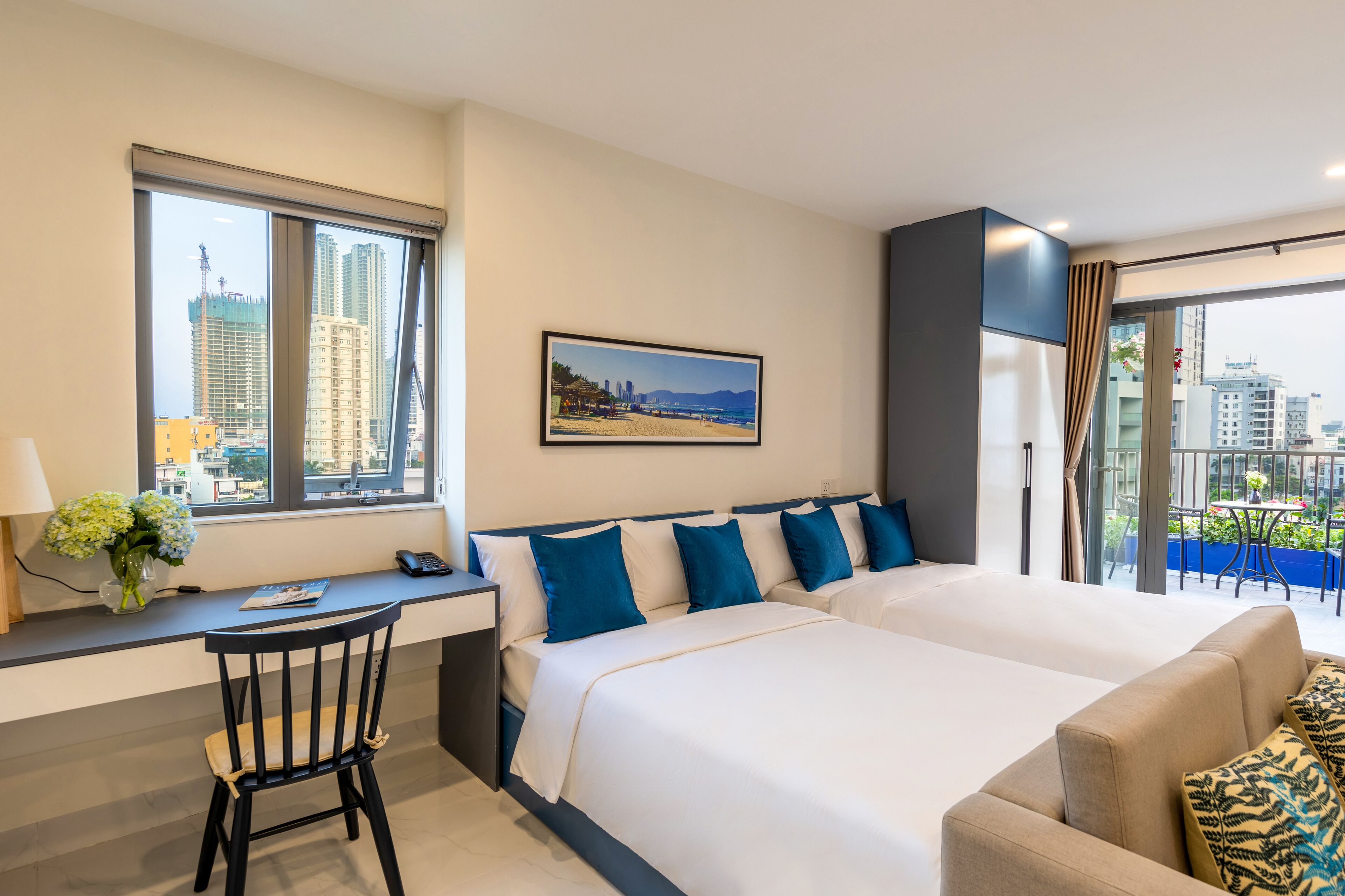 A hotel room with a large bed, a desk with a chair, a window with a view of the city, and a balcony with outdoor seating.