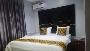 Een net opgemaakt bed met een wit-gele sprei, een donker hoofdbord met gouden accenten, en een airconditioning erboven.