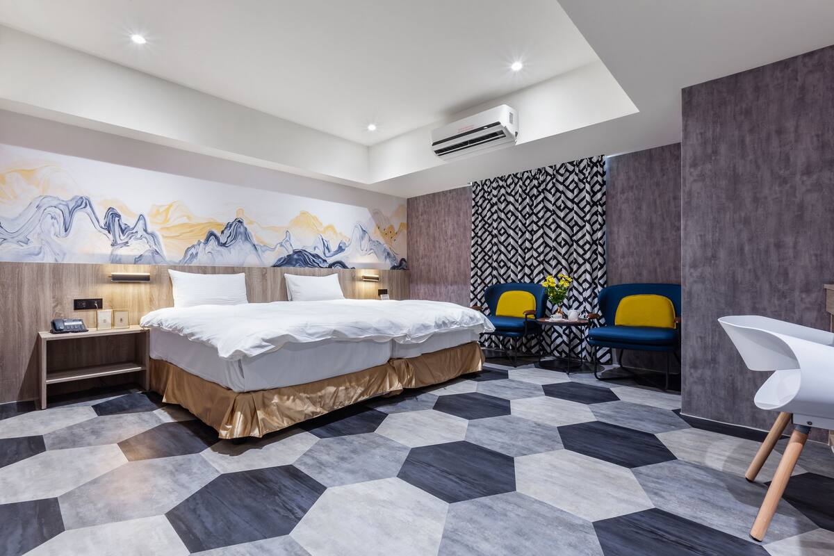 A modern hotel room with a large bed, a desk with a chair, a small table with a vase, and a wall with a mountain painting.