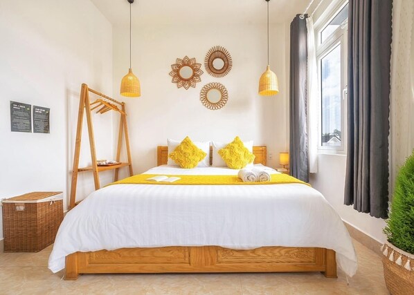 A neatly made bed with a wooden frame, a yellow throw blanket, and two pillows. There are two hanging lamps, a window with curtains, and a small plant on the windowsill.
