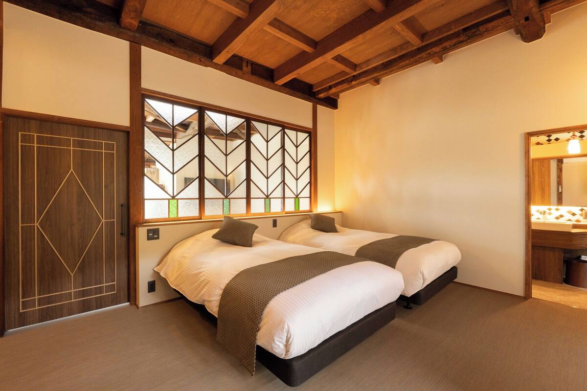 Ein Schlafzimmer mit zwei Betten, einer Holzbalkendecke, einem großen Fenster mit geometrischem Muster und einer Holztür.