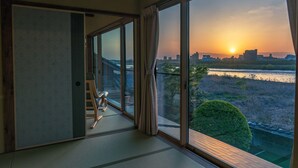 Een kamer met een grote schuifdeur die uitkomt op een balkon met uitzicht op een zonsondergang boven een stadshemelbeeld en een rivier.
