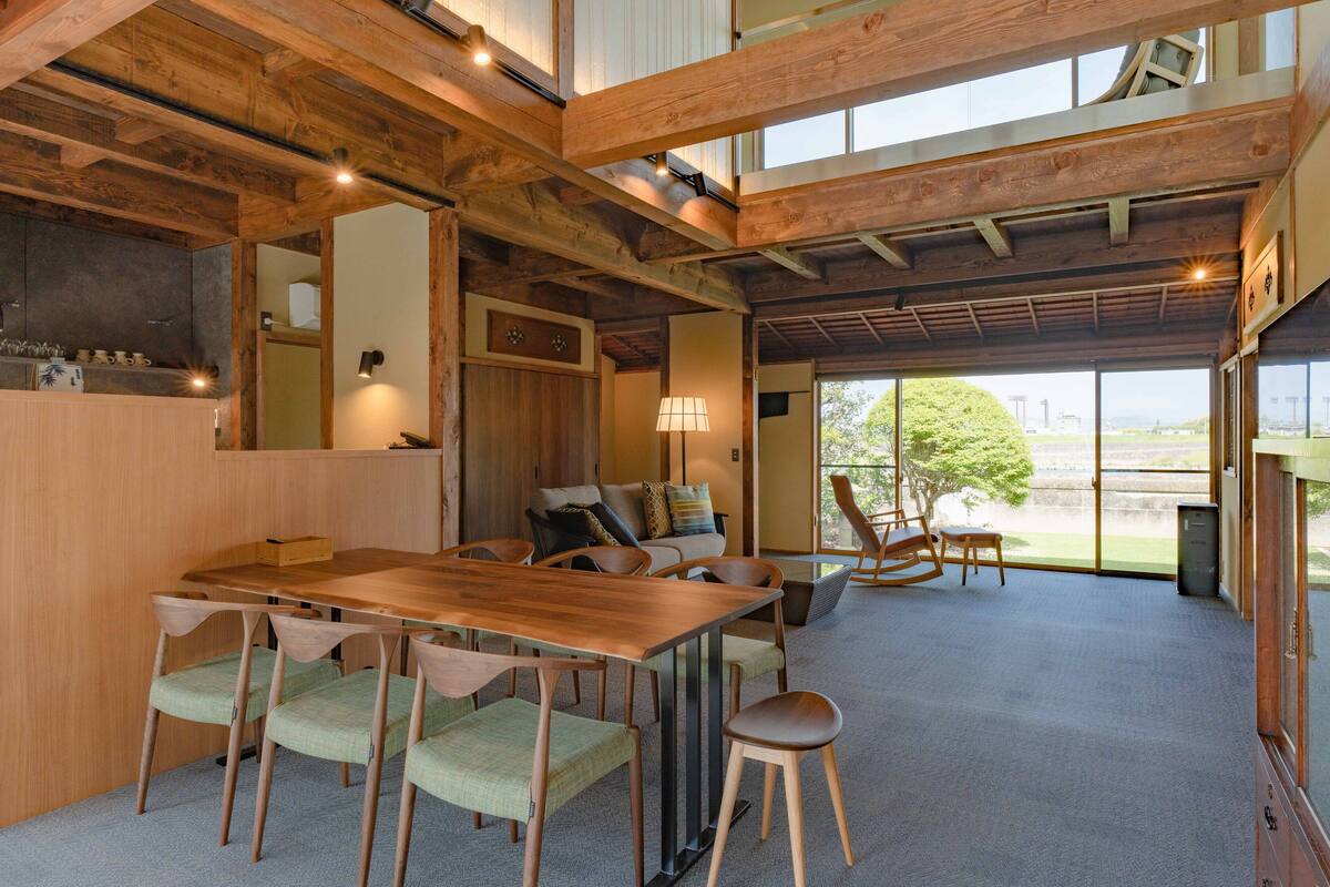 Ein geräumiges Zimmer mit einem Holzespeisetisch, Stühlen und einem Sofa. Eine Lampe, ein kleines Schränkchen und ein Blick ins Freie durch große Fenster sind ebenfalls vorhanden.