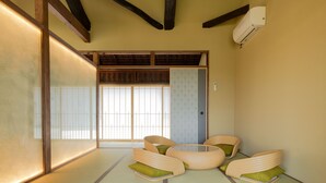 A traditional Japanese room with tatami flooring, low wooden beams, a sliding door, and a small round table with cushions.