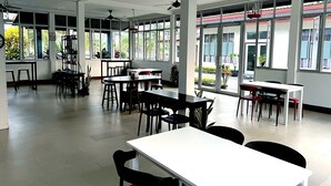 Un espace intérieur spacieux, doté de nombreuses tables et chaises, de grandes fenêtres laissant entrer la lumière naturelle et d’un ventilateur de plafond.