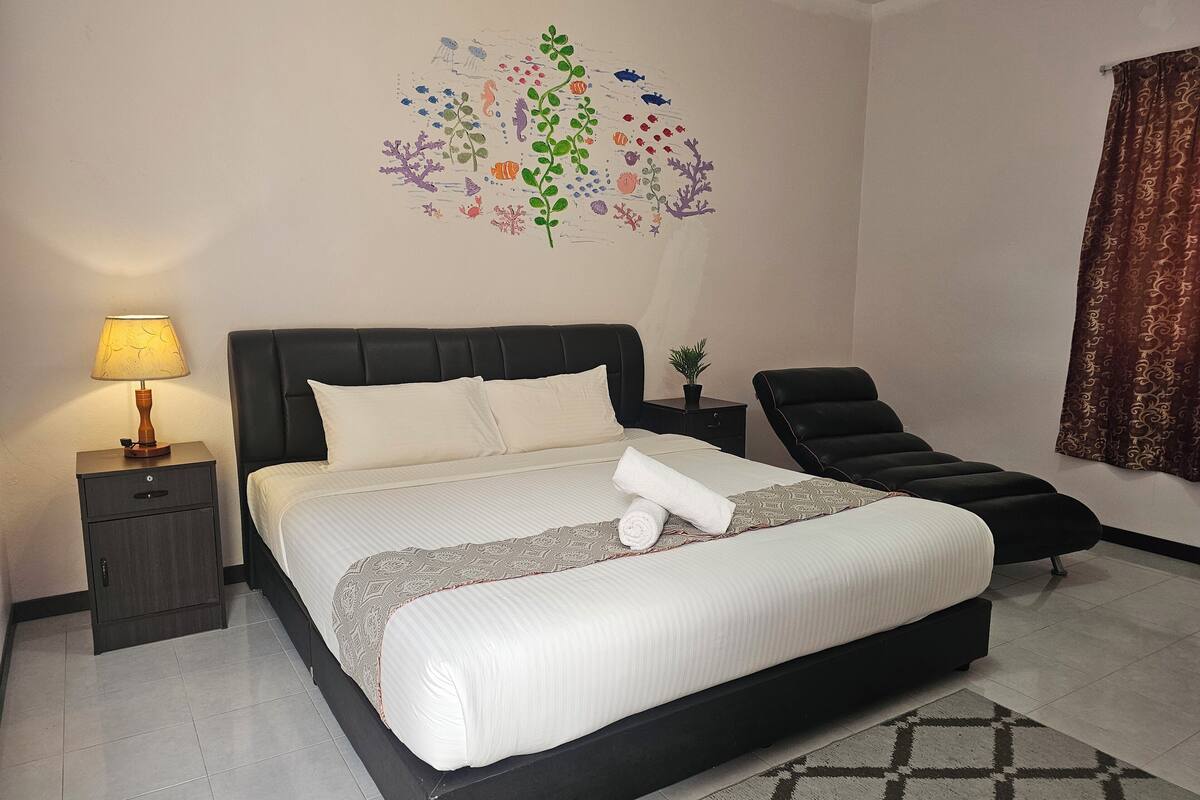 Un lit bien fait, avec une couette blanche et des oreillers, une lampe de chevet, une petite plante en pot, un fauteuil en cuir noir et une fresque décorative sur le mur.