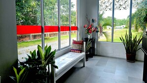 Une pièce avec un banc, des plantes en pot, une fenêtre avec un store rouge et une vue sur la verdure à l’extérieur.