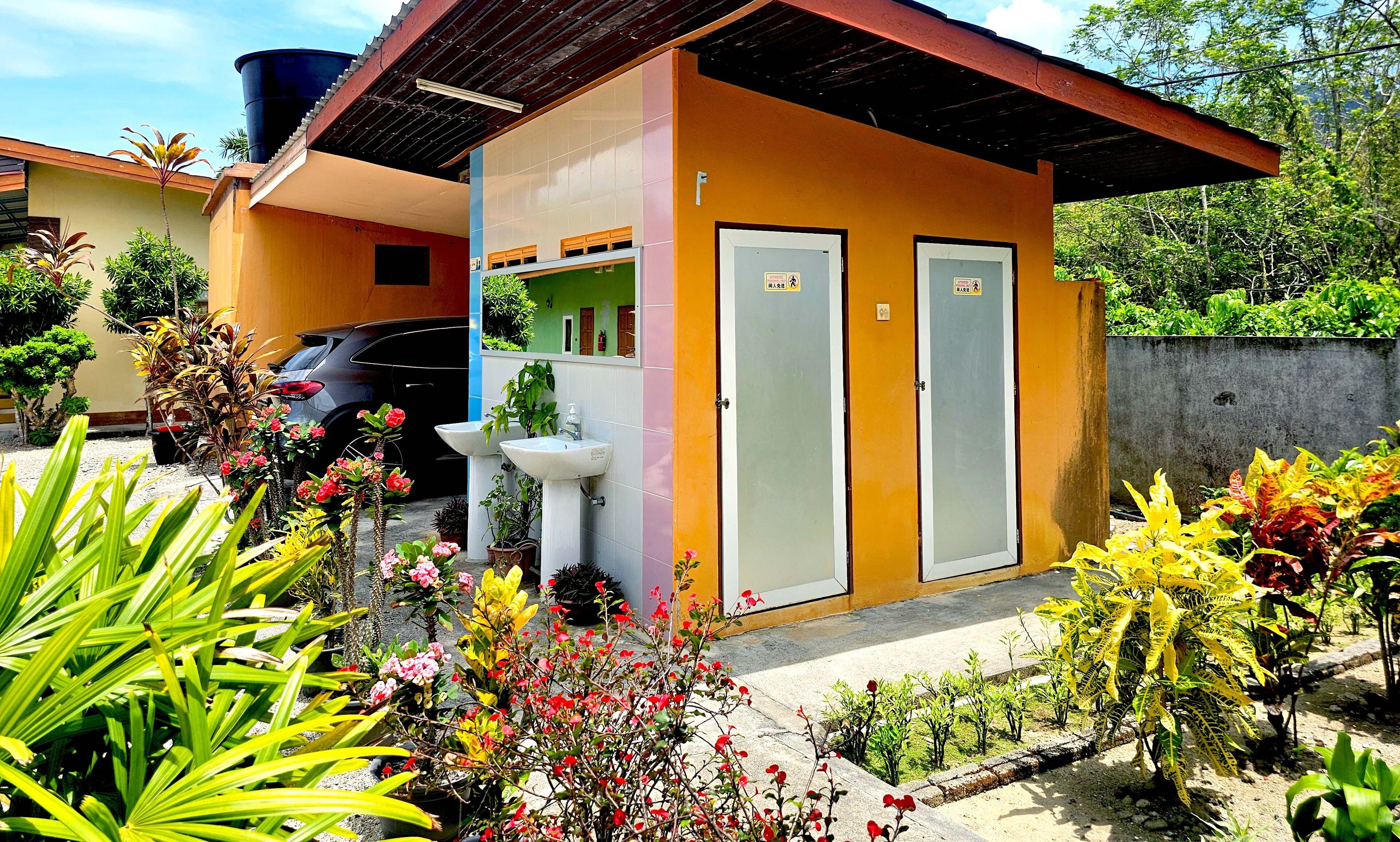 A small, two-story building with a yellow exterior, a black car parked in front, and a variety of potted plants and flowers around the entrance.