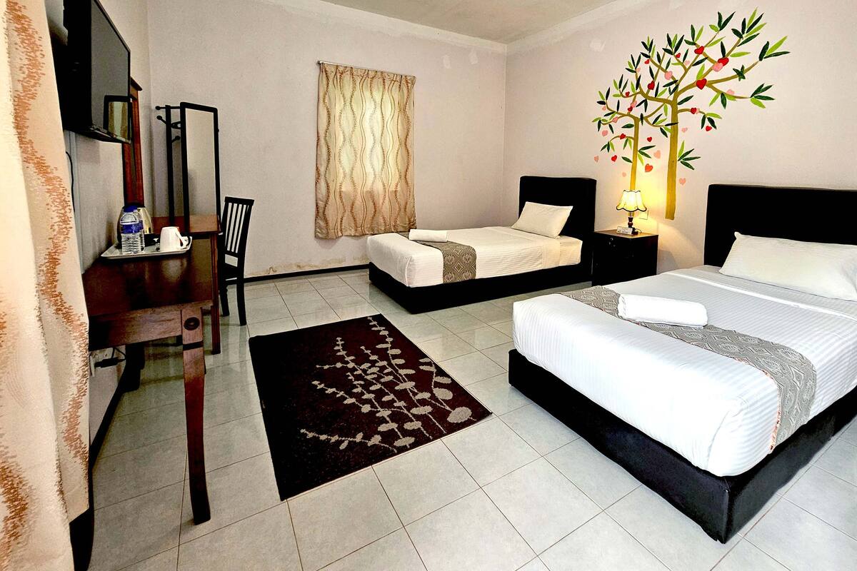 Une chambre d’hôtel comprenant un lit, un bureau avec une chaise, une petite table avec une bouteille et des verres, une fenêtre avec des rideaux et une décoration murale représentant un arbre orné de cœurs.