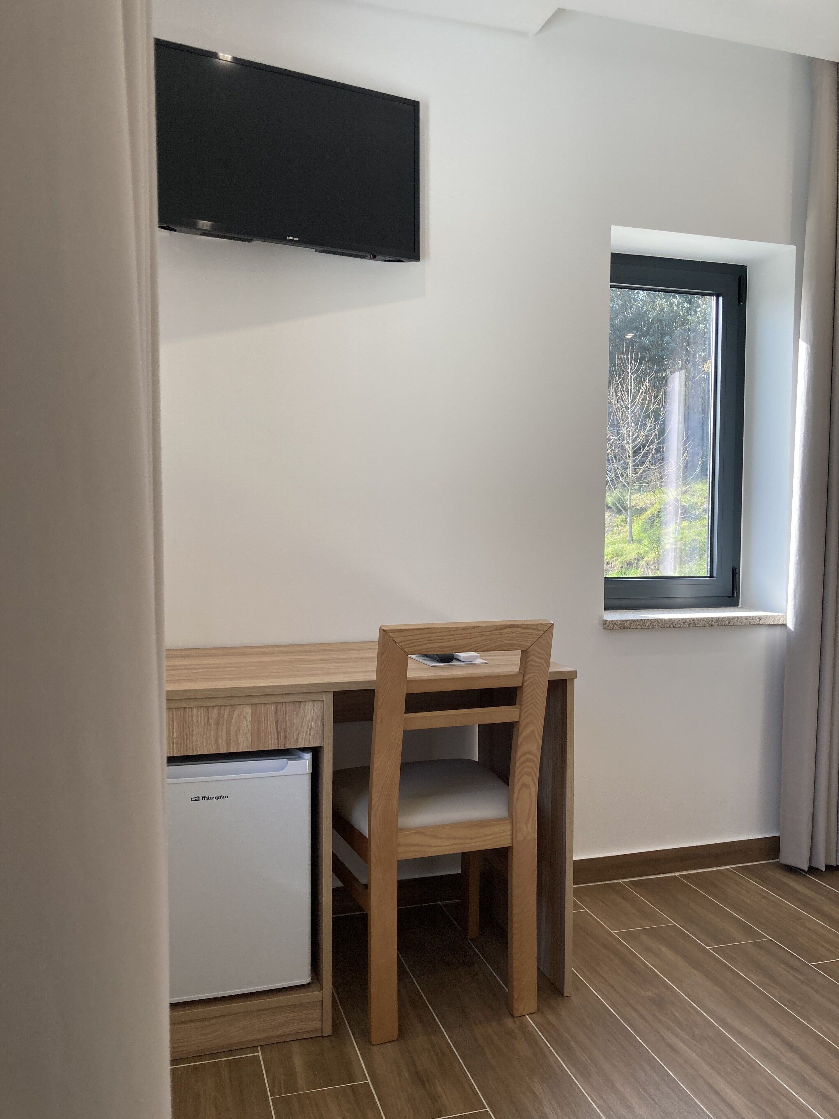 A compact room with a wooden desk, a chair, a flat-screen TV mounted on the wall, a small white refrigerator, and a window with a view of trees.