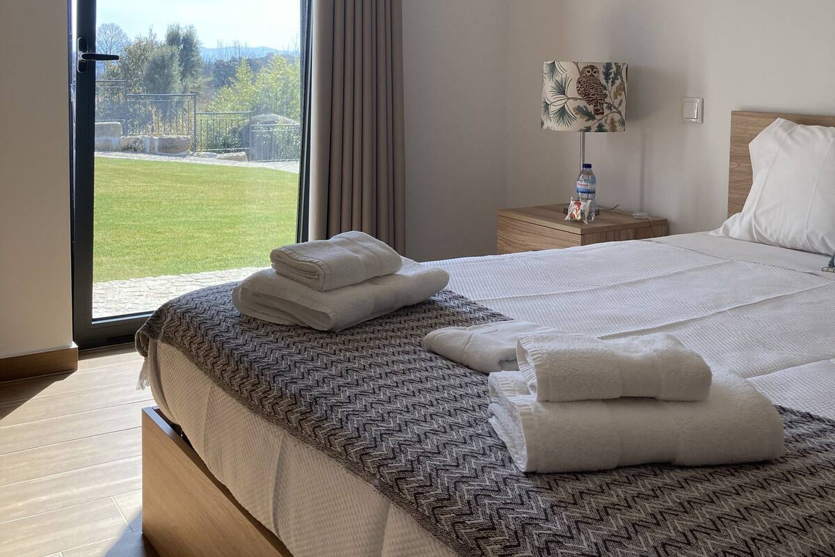 A hotel room with a large bed, a nightstand, a lamp, and a view of a grassy area through a sliding door.