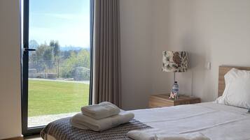 A hotel room with a large bed, a nightstand, a lamp, and a view of a grassy area through a sliding door.