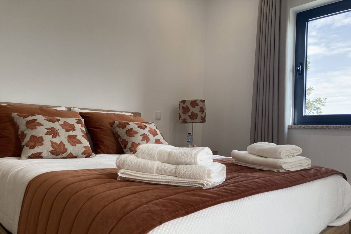A neatly made bed with a brown headboard, white bedding, and a brown leaf-patterned pillow. There are folded white towels on the bed and a water bottle on the nightstand.