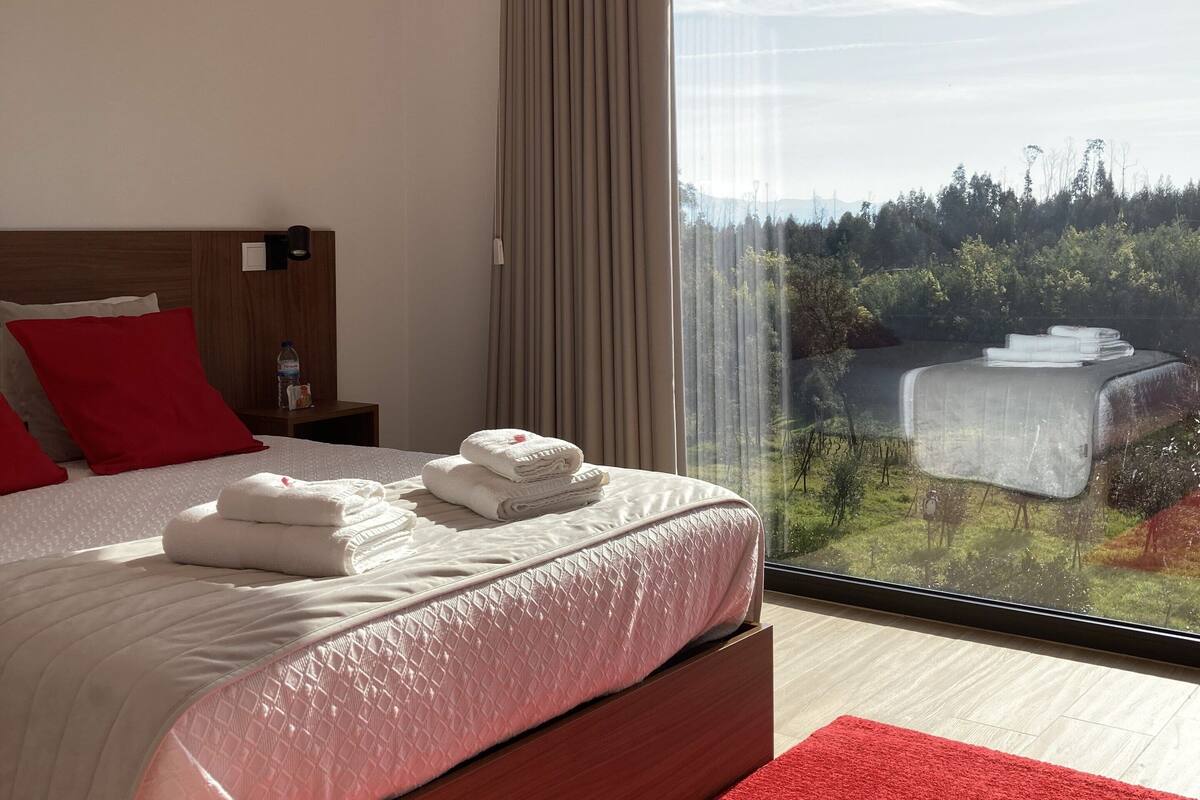A modern hotel room with a large bed, red pillows, a wooden headboard, a nightstand, and a view of a landscape through a large window.
