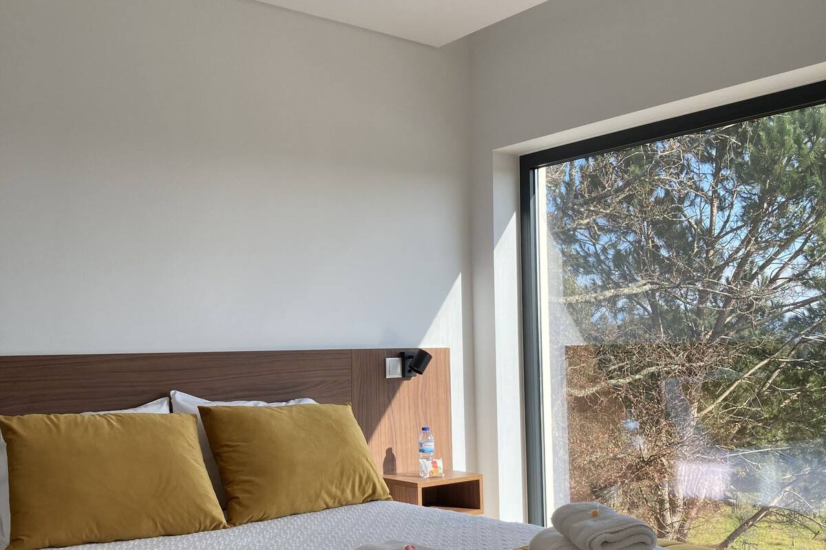 A modern bedroom with a large bed, a wooden headboard, a nightstand with a water bottle, and a view of trees through a large window.