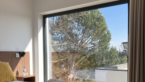 Une chambre moderne avec un grand lit, une table de chevet et une vue sur les arbres par une grande fenêtre.
