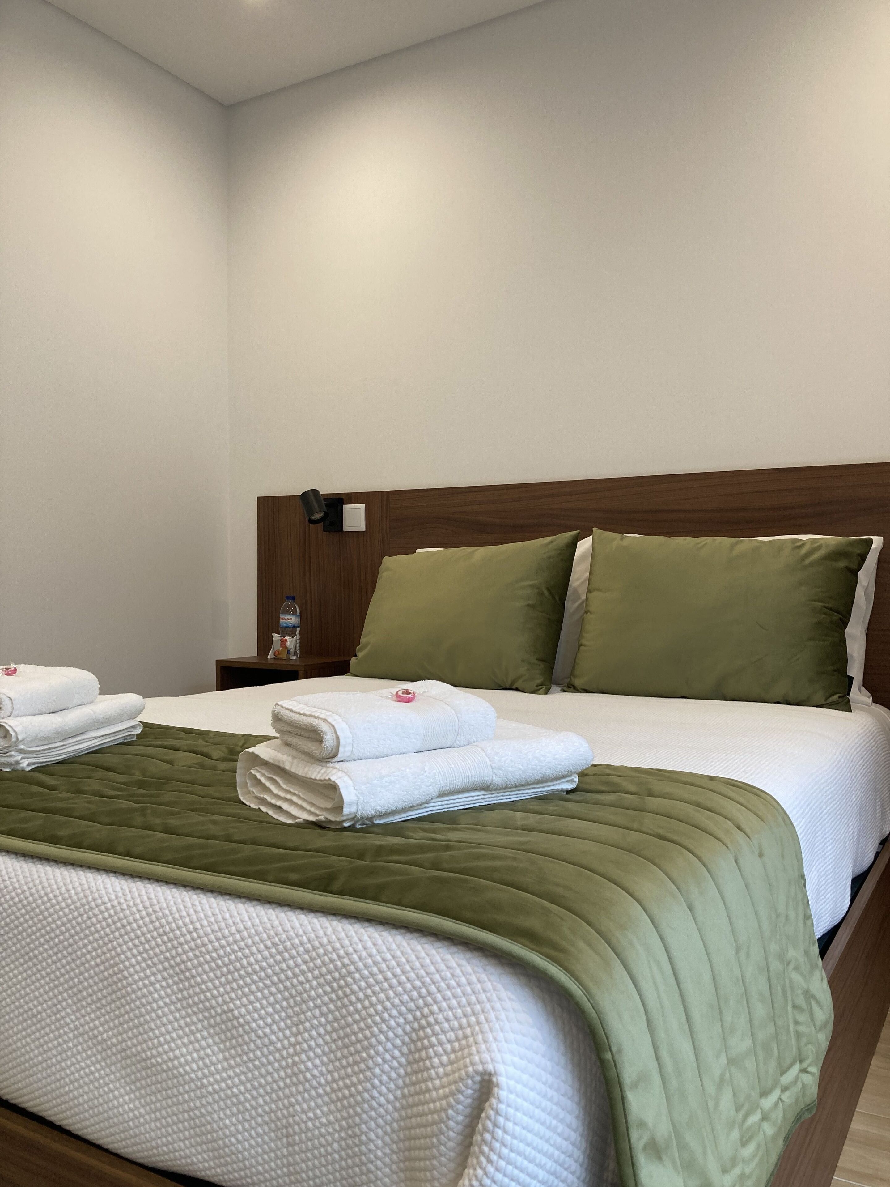 Quarto de hotel com cama bem arrumada, roupa de cama verde e branca, cabeceira de madeira e mesa de cabeceira com uma garrafa e um abajur.