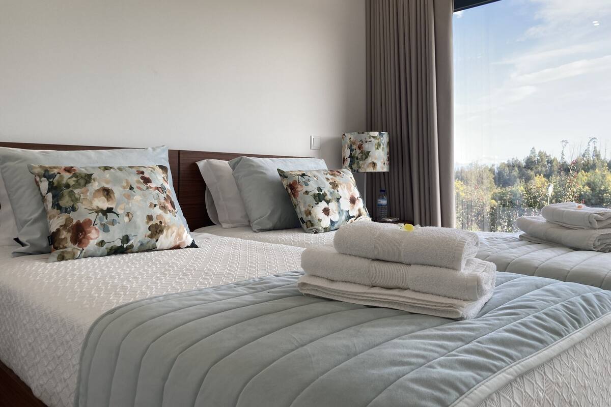A hotel room with two beds, floral-patterned pillows, a bedside lamp, and a view of trees through a window.