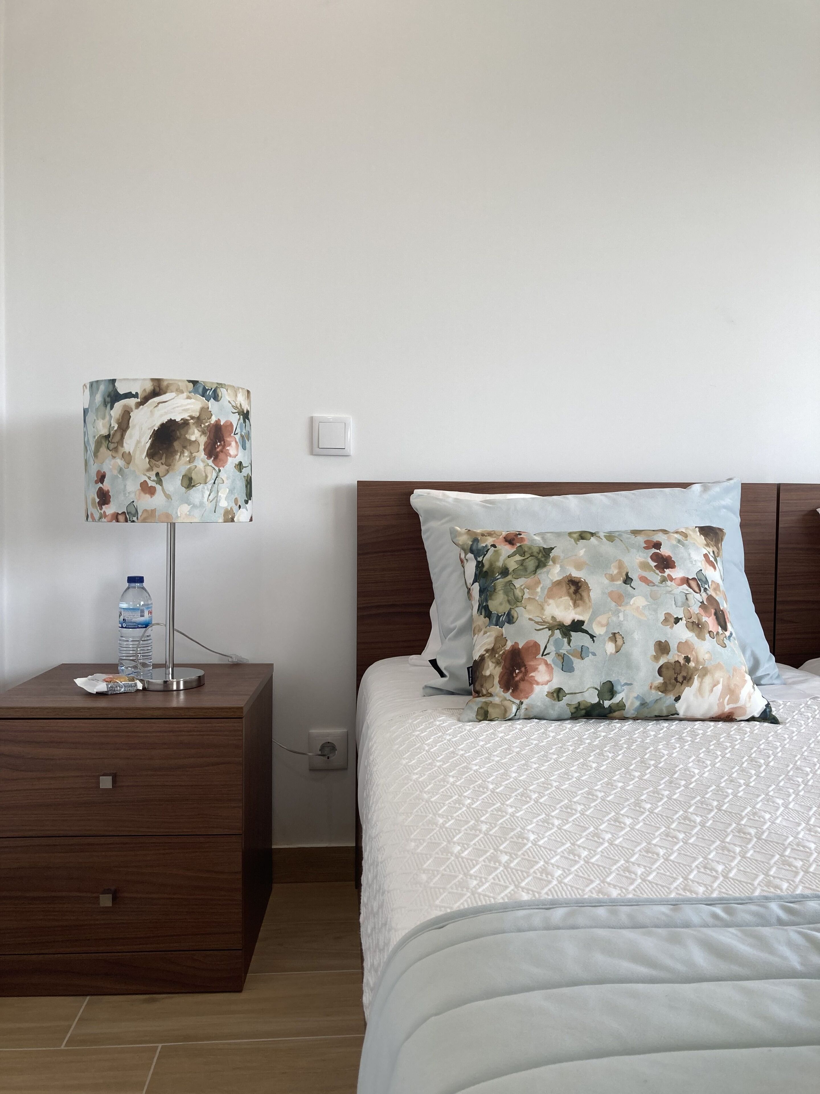 Una cama impecablemente hecha con un cojín floral y una colcha blanca, una mesita de noche con una lámpara y una botella de agua, y un tocador de madera con cajones.