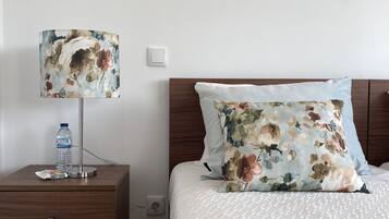 Una cama impecablemente hecha con un cojín floral y una colcha blanca, una mesita de noche con una lámpara y una botella de agua, y un tocador de madera con cajones.