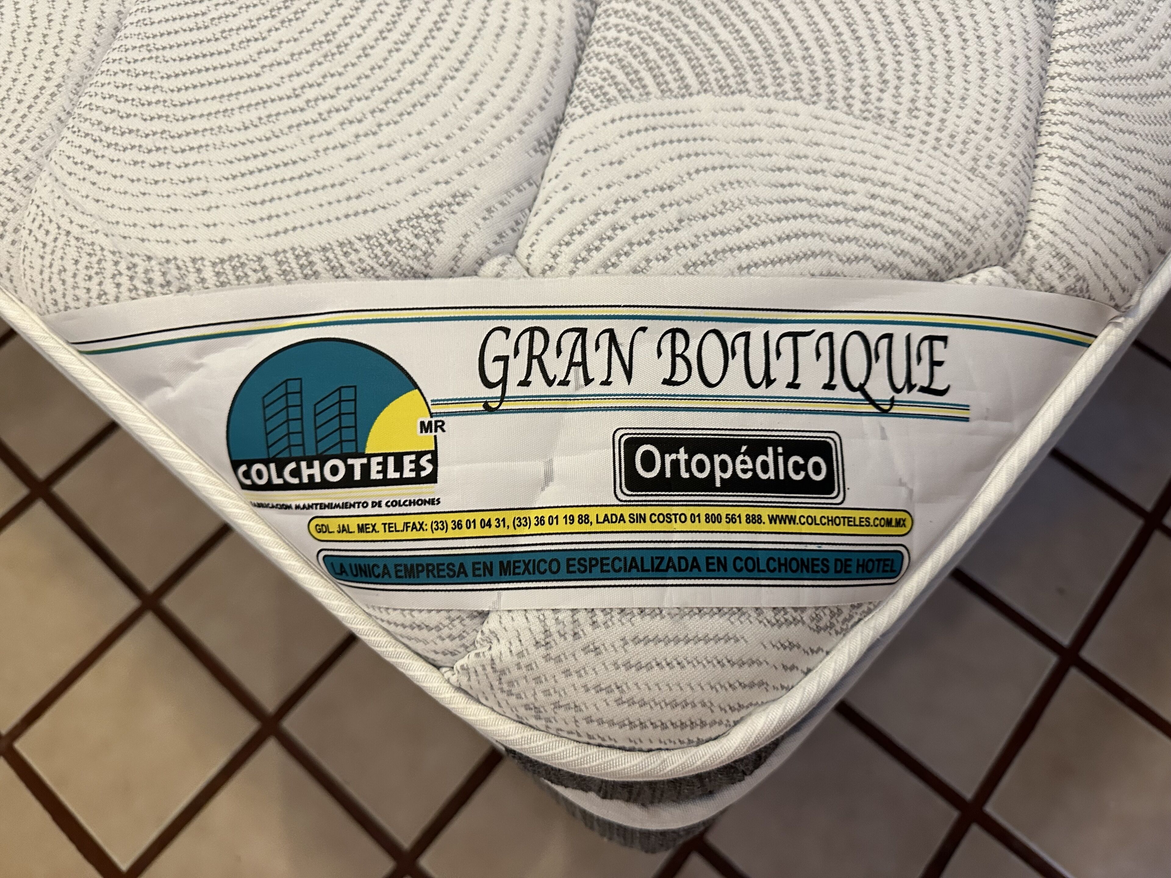 Quarto de hotel com uma cama coberta por um cobertor branco com a etiqueta que diz "GRAN BOUTIQUE" e "COLCHOTEALES".