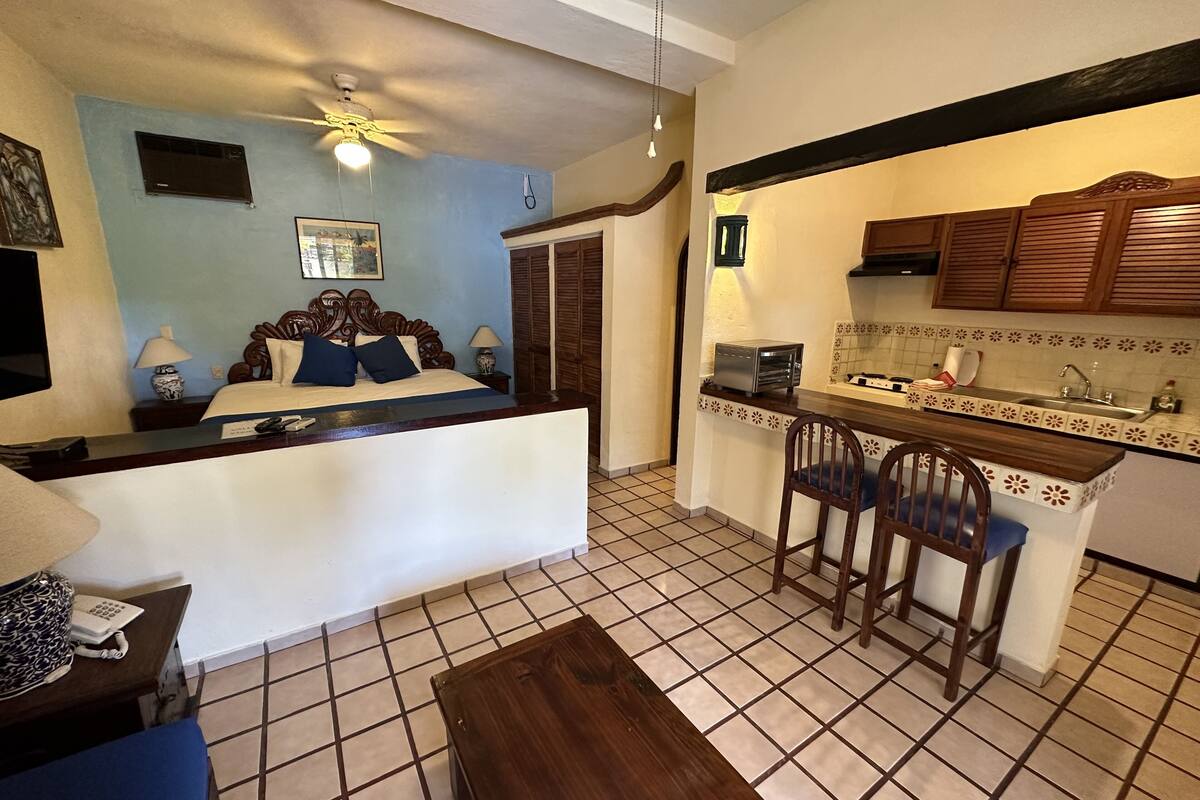 A compact hotel room with a kitchenette, a bed with blue bedding, a ceiling fan, and a tiled floor.