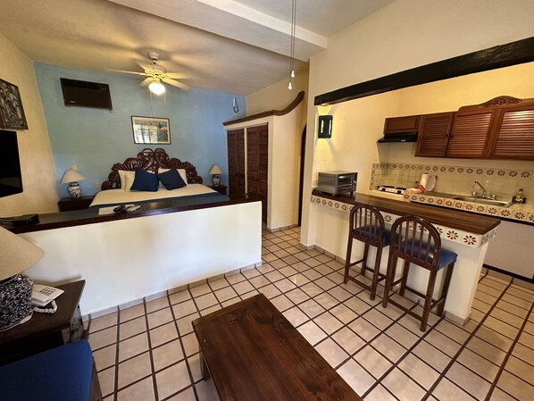 A compact hotel room with a kitchenette, a bed with blue bedding, a ceiling fan, and a tiled floor.