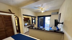 Habitación de hotel con cama, escritorio con computadora, ventilador de techo y vista a la cocina y al exterior.