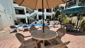 Un área junto a la piscina con sillones de descanso y una mesa bajo una sombrilla grande.