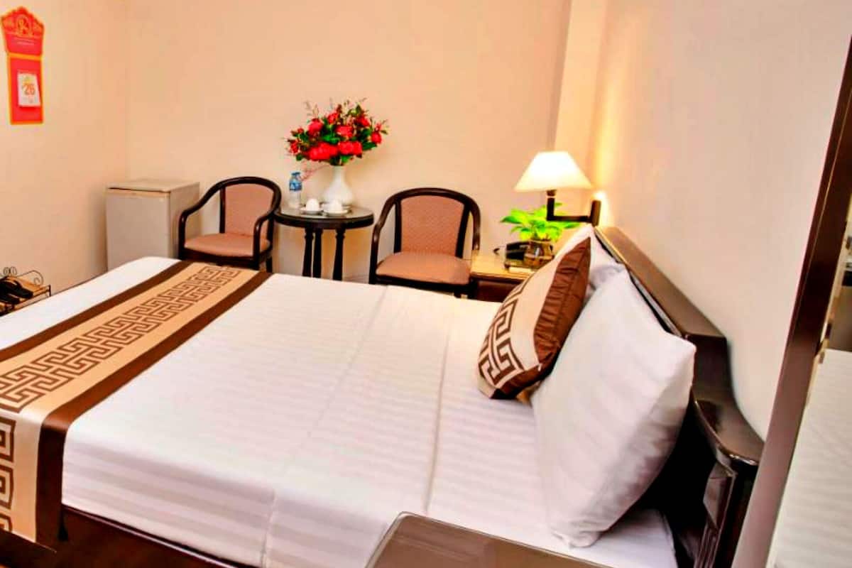 A hotel room with a bed, two chairs, a small table with a vase of flowers, a clock, and a lamp.