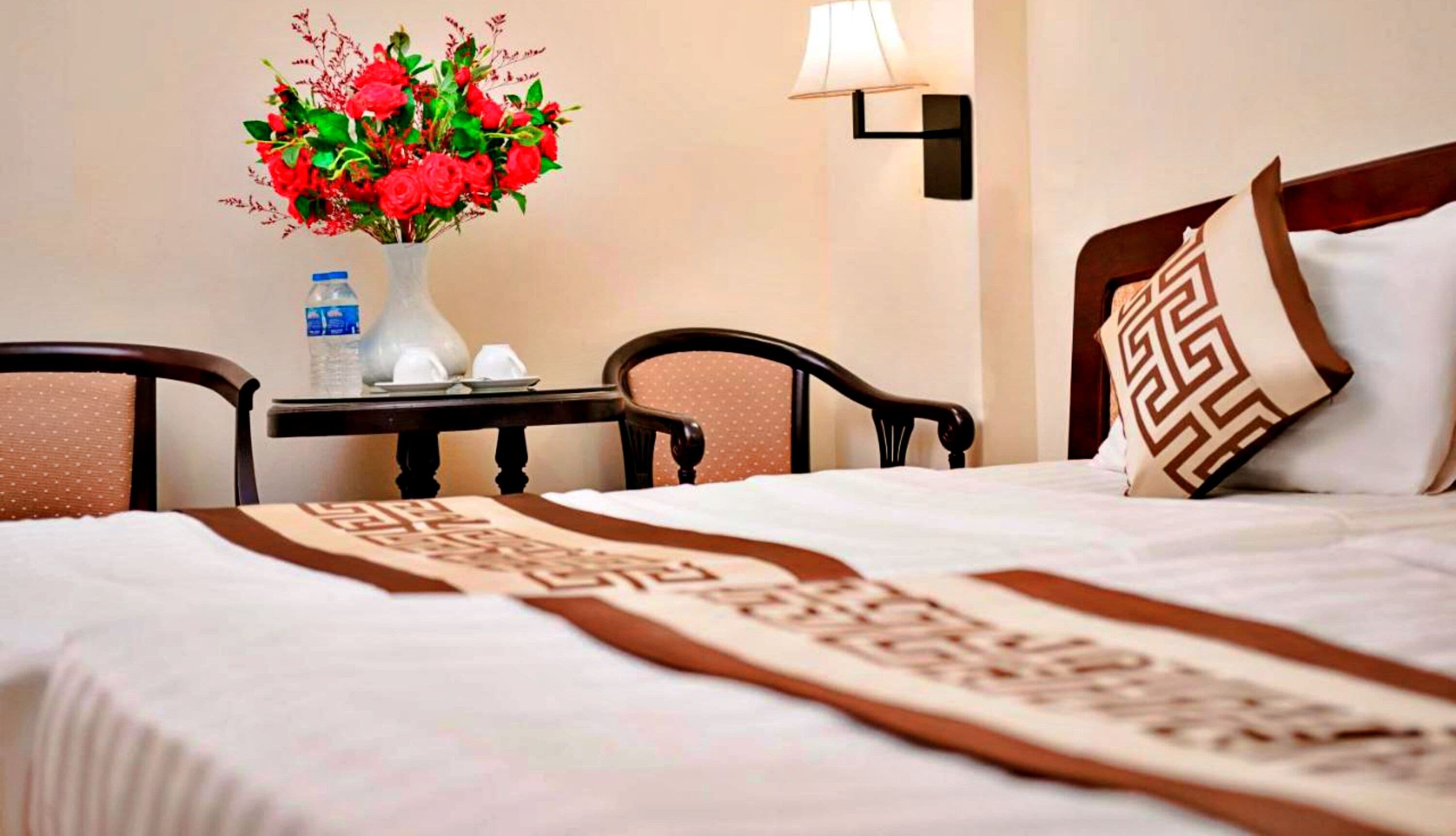 A hotel room with a bed, a nightstand, a vase of flowers, and a lamp.