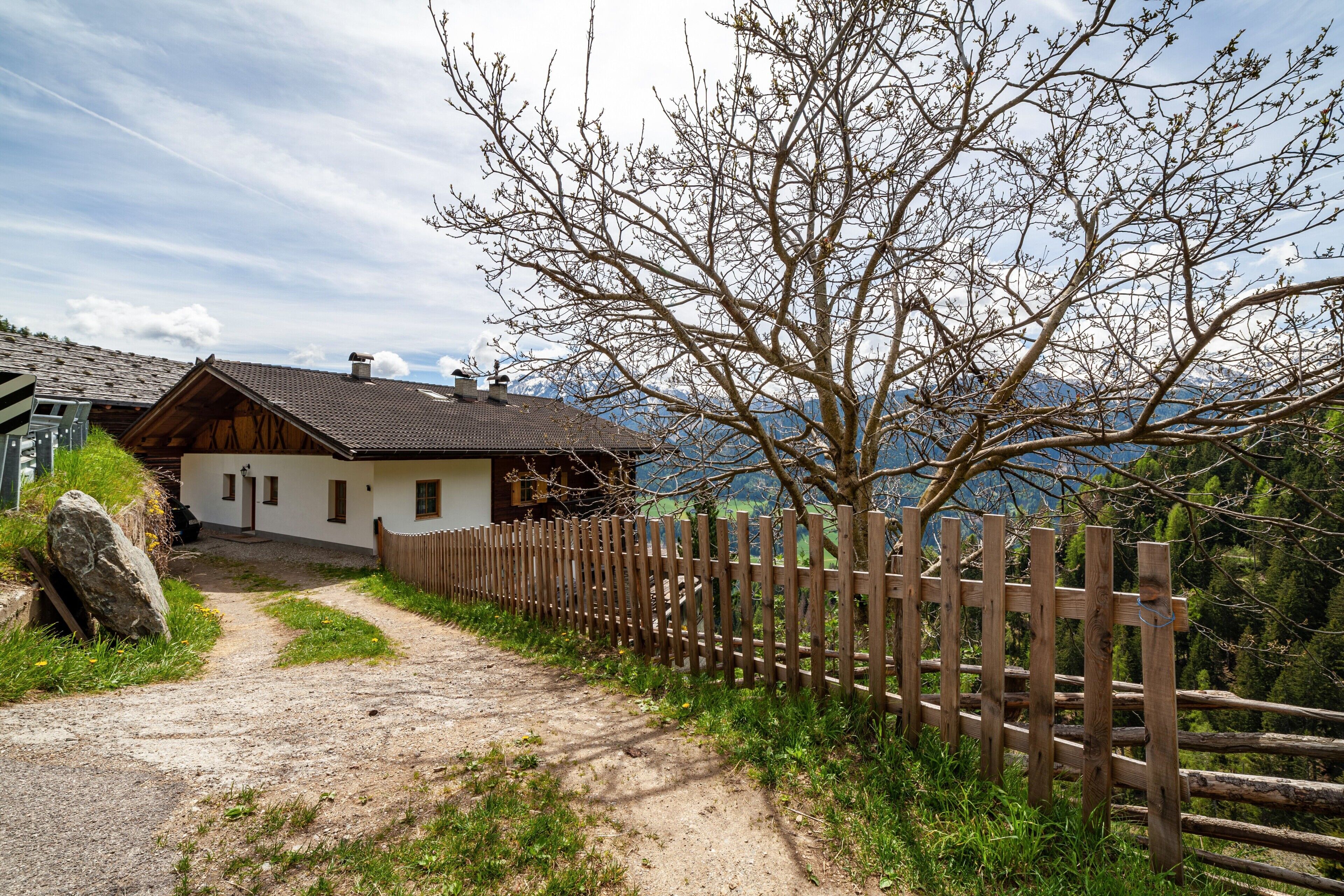 Una casa rustica con un sentiero sterrato che vi conduce, circondata da una recinzione in legno e un grande albero.