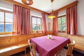 Une pièce avec un banc en bois, une table recouverte d’une nappe rose et de grandes fenêtres avec des rideaux.