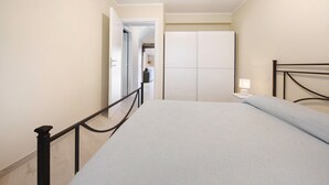 Una habitación de hotel con cama, mesita de noche, lámpara, armario y puerta que da a otra estancia.