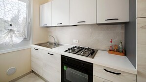 Una cocina con gabinetes blancos, una estufa a gas, un fregadero y una ventana con cortina decorativa.