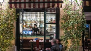 Un área de bar al aire libre con un toldo a rayas, estantes con botellas y un espacio de asientos con mesas y sillas.