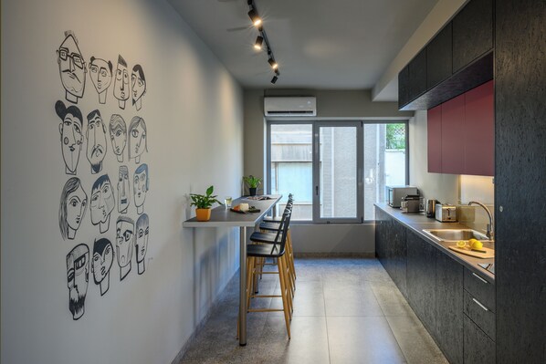 A modern kitchen with a long island, bar stools, a window with blinds, and a wall with black and white drawings.