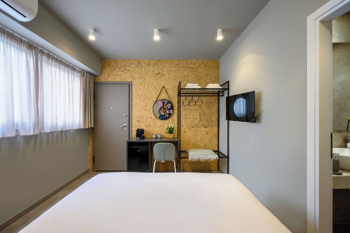 Habitación de hotel con cama, escritorio con silla, televisor montado en la pared y un pequeño rincón de descanso con un banco.