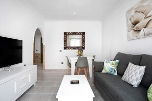 Un salon moderne doté d’un téléviseur à écran plat, d’une table basse blanche, d’un canapé gris et d’un coin repas avec une table et des chaises blanches.