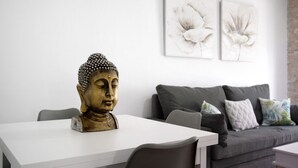 Una habitación con una mesa blanca, una estatua de Buda, un sofá gris con cojines y dos cuadros con motivos florales en la pared.