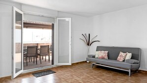 Una habitación con puerta corrediza de vidrio que da a un patio exterior, un sofá gris con cojines estampados y una planta alta.