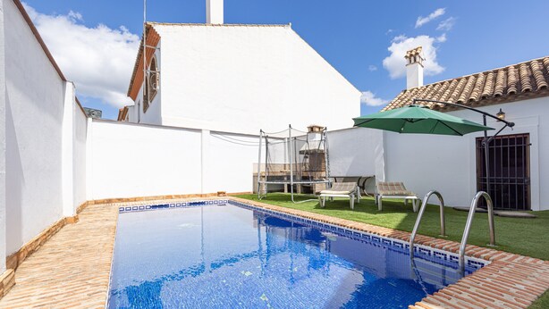 A swimming pool with a tiled edge, a green umbrella, and a white building with a tiled roof.