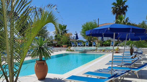 Un área de piscina con sillones de descanso y sombrillas, rodeada de palmeras y plantas en macetas.