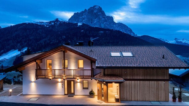 A two-story mountain lodge with a balcony, a separate wing, and a view of a mountain peak.