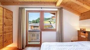 A bedroom with a wooden ceiling, a large window with curtains, a bed, and a wooden wardrobe.