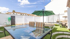 An outdoor patio with a glass table and striped chairs, a green umbrella, and a white wall with a door.