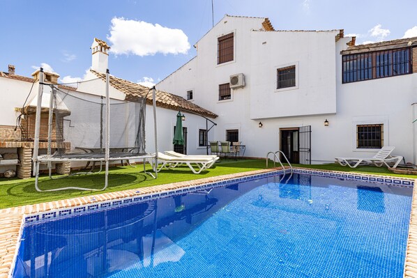 Una piscina con sillones de descanso, un patio y un edificio blanco con múltiples ventanas y techo de tejas.