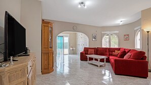 A spacious living room with a red sofa, a wooden cabinet, a television, and a large window. - Villa 'Casa Leuki' with Sea/Mountain Views, Pool, Garden, Wi-Fi & AC (Calpe)