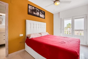 A bedroom with a white bed, red bedding, a ceiling fan, and a window with a view.